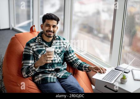 Portrait of nice-looking indian employee sitting on soft office chair with personal laptop on knees and smiling at camera. Imposing dark haired man taking short break after intensive working day. Stock Photo