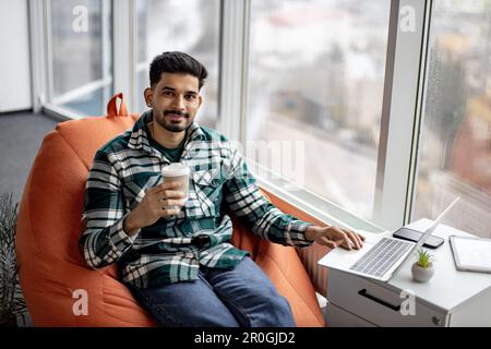 Portrait of nice-looking indian employee sitting on soft office chair with personal laptop on knees and smiling at camera. Imposing dark haired man taking short break after intensive working day. Stock Photo