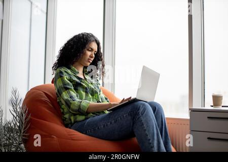 Attractive african american lady in casual apparel typing on portable computer while working remotely in office interior. Confident self-employed professional searching information for presentation. Stock Photo