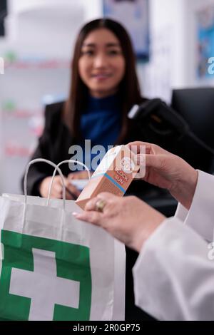 Pharmaceutical employee putting sunscreen in customer shopping bag at pharmacy paydesk. Drugstore cashier holding dermatologic product, packing client purchase in paper package close up Stock Photo