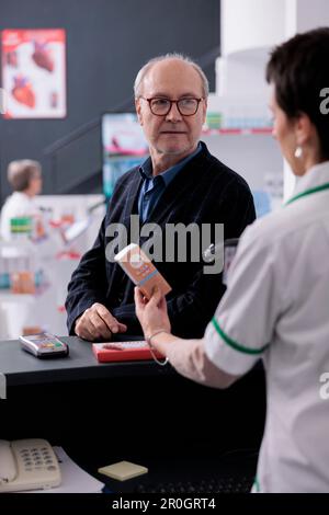 Old man in glasses waiting for drugstore cashier scanning product to pay. Young woman pharmaceutical worker using scanner on sunblock lotion barcode, selling medications to client Stock Photo