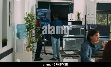 Diverse team of system developers analyzing code on big tv screen, checking errors in artificial intelligence project. Programmers working on machine learning software, developing new interface. Stock Photo