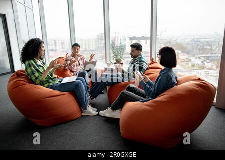 Young team of four multiethnic colleagues having conversation while seating on pouf chairs in open office. Active fellow workers in casual wears discussing business issues using digital devices. Stock Photo