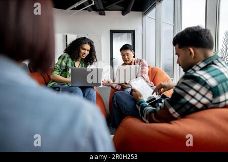 Focus on cheerful corporate people in jeans working on laptops while teammates using tablet in coworking space. Initiative multiethnic freelancers sharing content while making internet research. Stock Photo