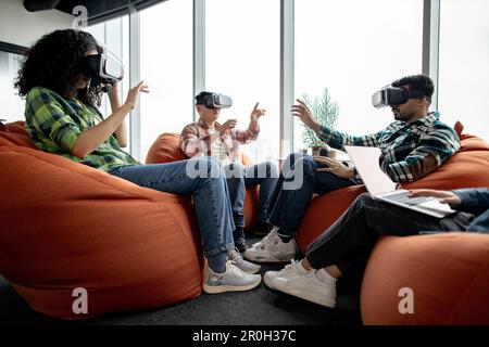 Multiethnic professionals in street style clothes using VR glasses while female colleague typing on laptop in creative workspace. Civil engineers finding building plan anomalies via modern technology. Stock Photo