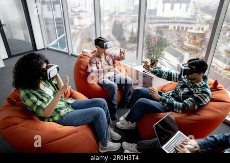 Multiethnic professionals in street style clothes using VR glasses while female colleague typing on laptop in creative workspace. Civil engineers finding building plan anomalies via modern technology. Stock Photo