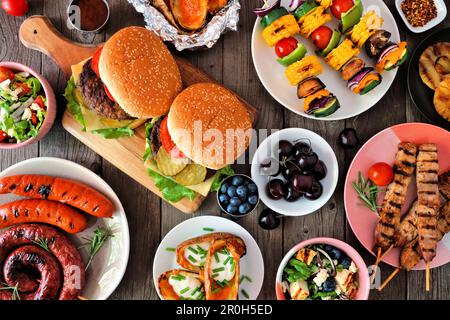 Summer BBQ or picnic food table scene. Selection of burgers, grilled meat, vegetables, fruits, salad and potatoes. Above view on a dark wood backgroun Stock Photo