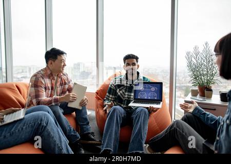 Close up of arabian male keeping portable computer while multiethnic team members taking seats on terracotta poufs. Automotive engineers generating creative ideas of car systems in office interior. Stock Photo