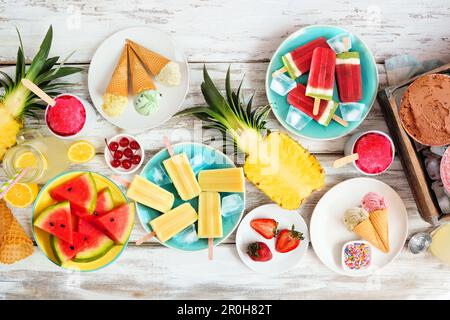Cool summer foods table scene. Collection of ice cream, popsicles and fruit. Overhead view on a rustic white wood background. Stock Photo
