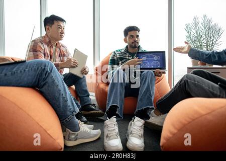Close up of arabian male keeping portable computer while multiethnic team members taking seats on terracotta poufs. Automotive engineers generating creative ideas of car systems in office interior. Stock Photo