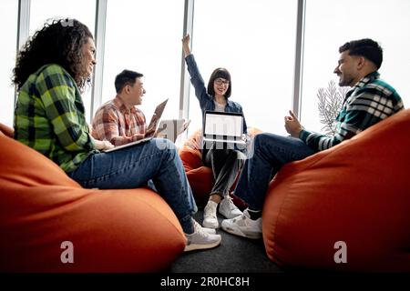 Beautiful caucasian lady in glasses raising right hand up while holding computer with sketches on screen. Multiracial designers celebrating successful construction plan while staying in workspace. Stock Photo