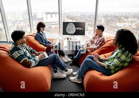 Full length view of diverse professionals working on gadgets while staying in open office with AI presentation on display. Efficient scientists developing online customer support system in office. Stock Photo