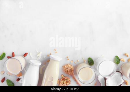 Vegan, plant based, non dairy milk bottom border. Variety of types in milk bottles and glasses with scattered ingredients. Top view over a white marbl Stock Photo