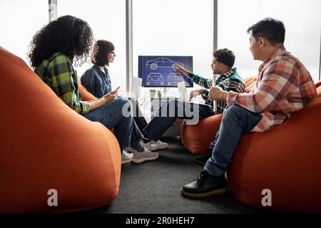 Diverse team of professionals utilizing smart gadgets while being busy at meeting in coworking space with soft poufs. Automotive engineers in casual wear analyzing digital data on display in office. Stock Photo
