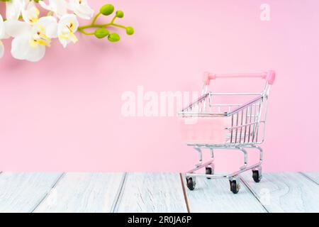Empty toy metal supermarket shopping cart on wooden floor with white orchid flower. Stock Photo