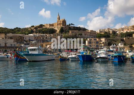 Traditional fishing boats in harbor and Our Lady of Lourdes Church on hillside, Mgarr, Gozo, Malta Stock Photo