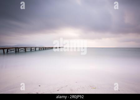 Long exposure of a wooden pier on Playa de Muro beach in the morning mood. The sky is overcast and fog lies over the Bay of Alcudia. Alcudia, Mallorca Stock Photo