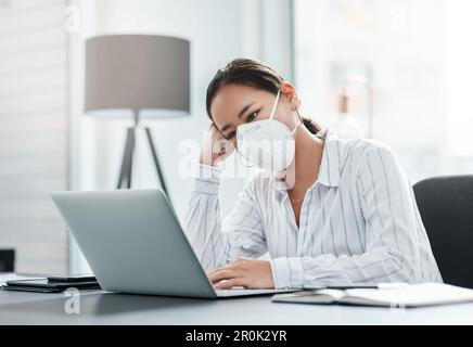 The world isnt well and everyone is feeling it. a masked young businesswoman looking unhappy while working at her desk in a modern office. Stock Photo