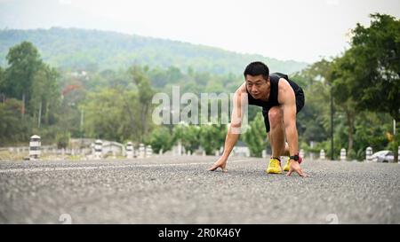 Determined and fit mature Asian man in sportswear is on the road with a starting position, preparing to run. Stock Photo
