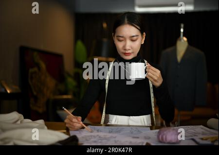 Concentrated and professional millennial Asian female fashion designer sipping coffee and working on her new collection in her studio. Stock Photo