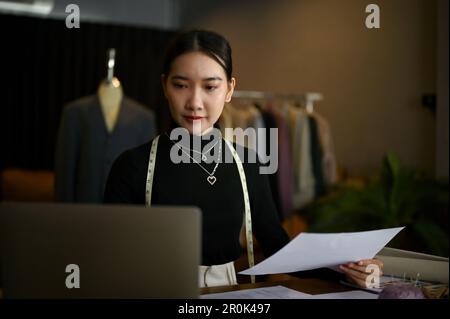 Focused and professional millennial Asian female fashion designer or tailor using laptop and working on her new collection in her studio. Stock Photo