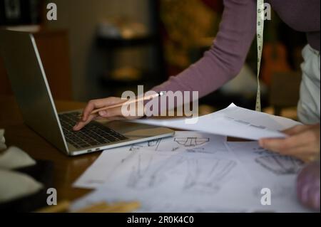 Close-up image of a professional female fashion designer or tailor using laptop computer and working on her new costume collection design in a studio. Stock Photo
