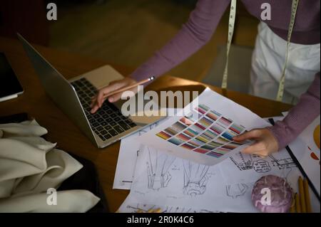 Close-up image of a professional female fashion designer using laptop and choosing color from color swatches chart, working in her studio. Stock Photo
