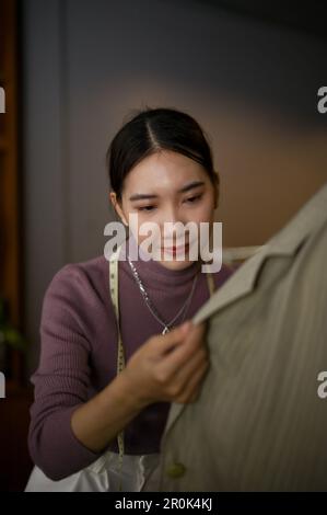 Portrait, A beautiful young Asian female fashion designer checking fabric on her new sample on a mannequin in her workshop studio. Stock Photo
