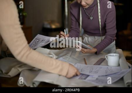 Close-up image of happy Asian female fashion designer checking new clothes design on tablet and working with her team in the studio. Stock Photo