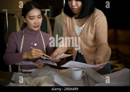 Two professional Asian female fashion designers or tailors are discussing and planning their new collection, and working in the studio together. Stock Photo