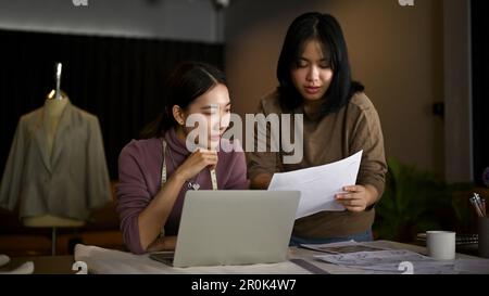 Two Asian female dressmakers or fashion designers are discussing their new collection's pattern and working in the studio together. Stock Photo
