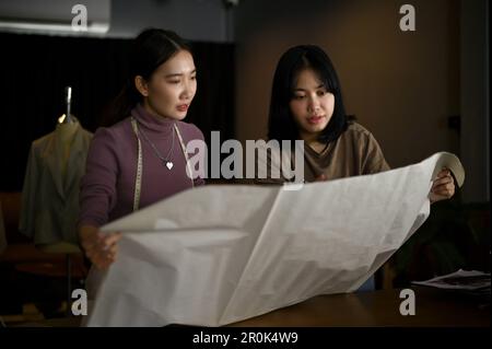 Two Asian female dressmakers or fashion designers are discussing, checking the quality of fabric for their new collection and working in the studio to Stock Photo