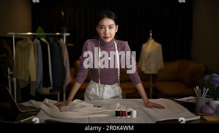 Portrait of a confident and successful millennial Asian female fashion designer or tailor standing at her desk in her studio. Stock Photo