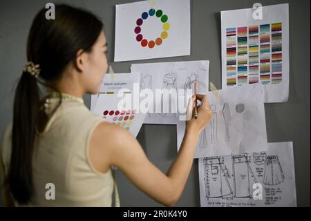 Focused young Asian female fashion designer checking model sketches on the wall, working in her creative studio. back view Stock Photo