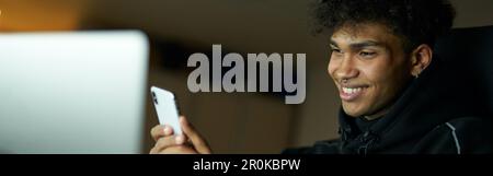 Distraction. Portrait of happy male student smiling and using smartphone while studying, working on laptop late at night Stock Photo