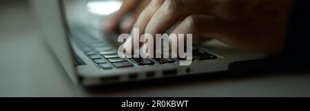 Creating process. Close up shot of hands of young guy typing, working on laptop, sitting at the table late at night Stock Photo