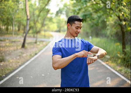 Handsome millennial Asian male runner in sportswear is checking time and running miles on his smart watch while running in a park. Stock Photo