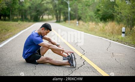 Athletic Asian man having muscle cramped during running, stretching his leg on the street in a park. Injured, leg pain Stock Photo