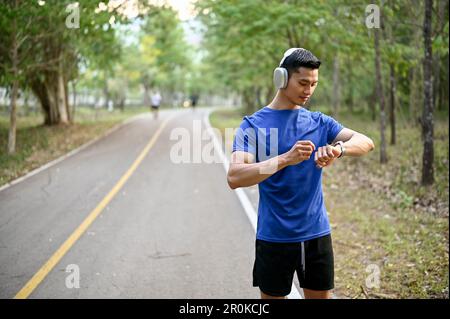 Handsome millennial Asian man wearing headphones, checking his heart rate and running miles on his smartwatch during running in a public park. Stock Photo