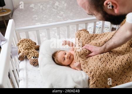 The father takes care of the newborn baby lying in the crib and looks at him. Stock Photo