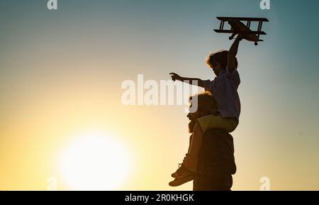 Sunset silhouette of Father carrying his son on shoulders. Boy child is sitting on daddy shoulder piggyback while the flight. Stock Photo