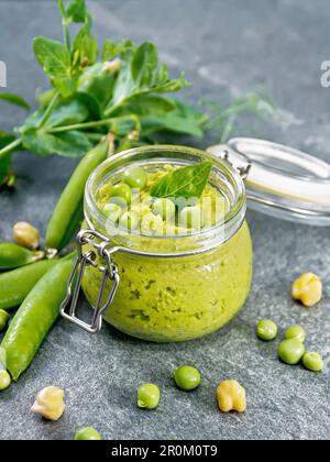 Hummus from green peas and chickpeas in a glass jar, pea pods on granite countertop background Stock Photo