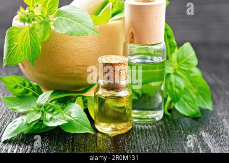 Oil with basil in two vials, mortar with a pestle and basil leaves on a dark wooden board background Stock Photo