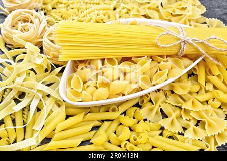 Pasta seashells and spaghetti in a bowl, pipe rigate, tagliatelle, fusilli, penne, farfalle, conquilla, trenette, rotini, pappardelle on black wooden Stock Photo