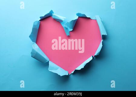 Torn heart shaped hole in light blue paper on pink background Stock Photo