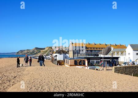 People walking along the pebble beach with views of the town and coastline, West Bay, Dorset, UK, Europe. Stock Photo