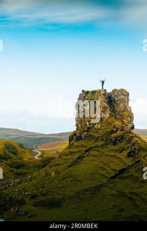 A person is standing atop a rocky mountain peak surrounded by lush green rolling hills in the background Stock Photo