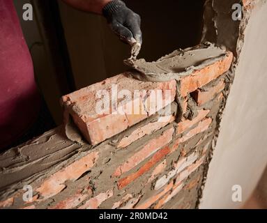 Close up of man hand in work glove laying brickwork in building under construction. Male worker applying cement mortar on bricks with trowel tool. Masonry construction concept. Stock Photo