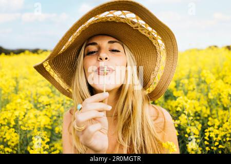 Beautiful young blonde cheerful woman, blowing dandelion seeds amidst a field of blooming yellow rapeseed flowers Stock Photo