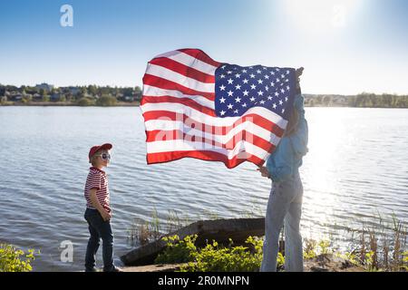 woman and child are standing on shore of lake holding an American flag waving in hands, beautifully illuminated by the sun. patriotic holiday, Indepen Stock Photo